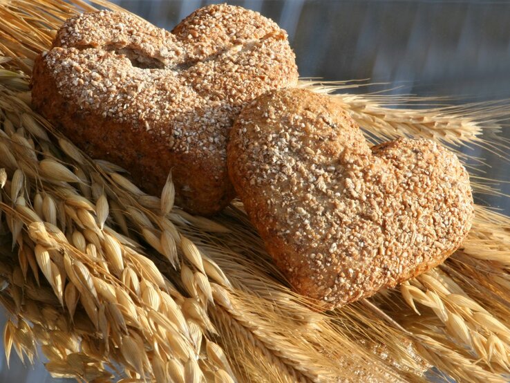 Zwei herzförmige Vollkornbrötchen, bestreut mit Sesamkörnern, liegen auf einem Bündel goldener Weizenähren. Die warme Beleuchtung betont die goldbraune Farbe des Gebäcks und das strahlende Gelb des Weizens, was eine Verbindung von frisch gebackenem Brot und seinem Ursprung im Kornfeld suggeriert. Die Komposition strahlt eine rustikale und gemütliche Atmosphäre aus.