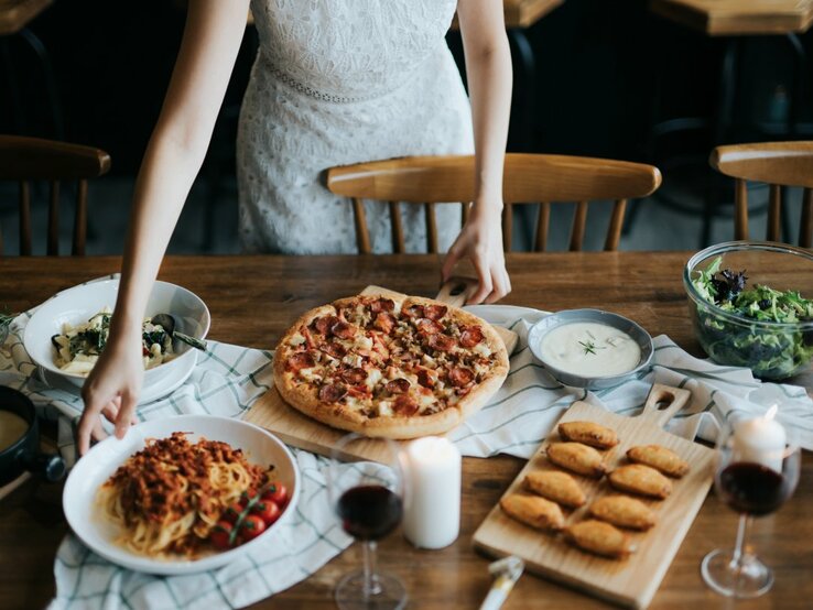 Eine Person in einem weißen Spitzenkleid bereitet am Tisch ein italienisches Essen vor. Auf dem Tisch befindet sich eine Vielfalt an Gerichten: eine Pizza mit reichlich Belag auf einem Holzbrett, ein Teller mit Spaghetti und Tomatensoße, ein Salat in einer Glaschüssel, eine Schüssel mit einer hellen, cremigen Suppe und eine Platte mit goldbraunen, gebackenen Teigtaschen. Zwei Gläser Rotwein und eine Kerze tragen zur gemütlichen Atmosphäre bei.
