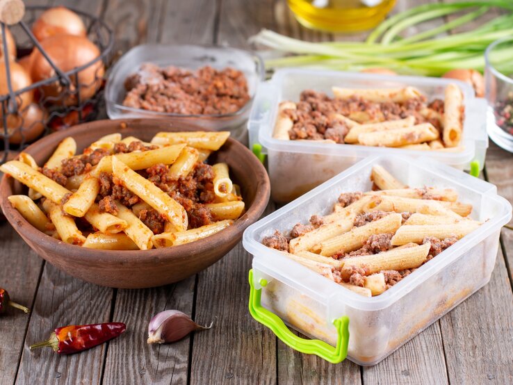 Das Bild zeigt eine hölzerne Schüssel gefüllt mit Pasta, die anscheinend mit einer Bolognese-Sauce gemischt ist, sowie mehrere Behälter mit der gleichen Pasta, die für die Lagerung oder das Einfrieren vorbereitet zu sein scheinen. Im Hintergrund sind einige Küchenzutaten und -utensilien sichtbar, darunter Zwiebeln, Knoblauch, Pfefferkörner, grüne Zwiebeln und Olivenöl, was auf eine frische Zubereitung der Mahlzeiten hinweist. Die Szene deutet auf das Konzept des Meal-Prepping hin, bei dem Mahlzeiten im Voraus zubereitet und dann für später gelagert werden.