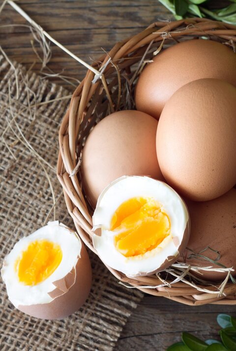 Die abgebildeten Gegenstände sind Eier, von denen eines aufgebrochen und offenbar hart gekocht ist, mit einem festen gelben Dotter. Sie liegen in einem Korb, der mit Stroh ausgelegt ist. Im Hintergrund sieht man eine rustikale Holzoberfläche und etwas Grünzeug, was eine natürliche, ländliche Atmosphäre vermittelt.