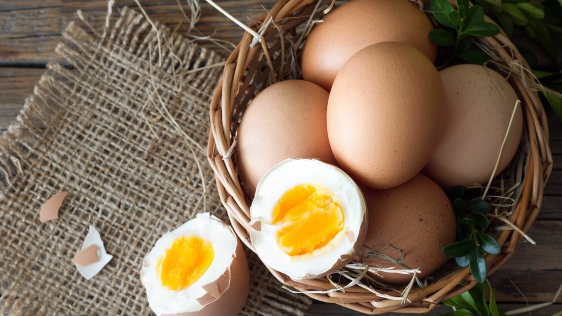 Die abgebildeten Gegenstände sind Eier, von denen eines aufgebrochen und offenbar hart gekocht ist, mit einem festen gelben Dotter. Sie liegen in einem Korb, der mit Stroh ausgelegt ist. Im Hintergrund sieht man eine rustikale Holzoberfläche und etwas Grünzeug, was eine natürliche, ländliche Atmosphäre vermittelt.