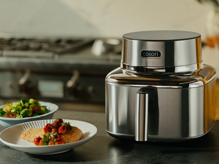 Elegante Heißluftfritteuse aus Edelstahl steht vor einer Kücheneinrichtung, daneben farbenfrohe Teller mit Gemüse und Fisch. | © Cosori
