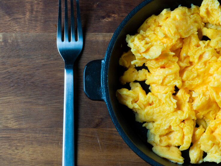 Cremiges Rührei in einer schwarzen Pfanne, angerichtet auf einem Holzbrett mit einer Gabel daneben.