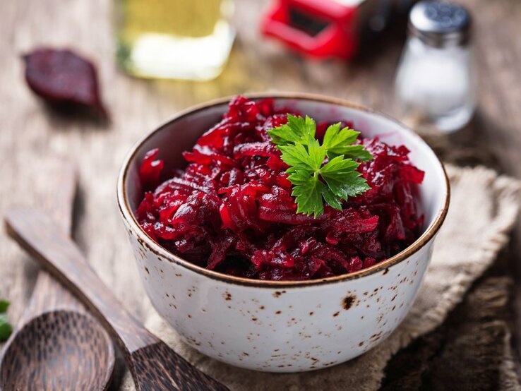 Das Bild zeigt eine Schüssel mit Rote-Bete-Salat, garniert mit einem Blatt Petersilie. Die Schüssel steht auf einem rustikalen Holztisch und im Hintergrund sieht man eine Ölflasche, eine Salzstreuer und vermutlich eine Pfeffermühle.