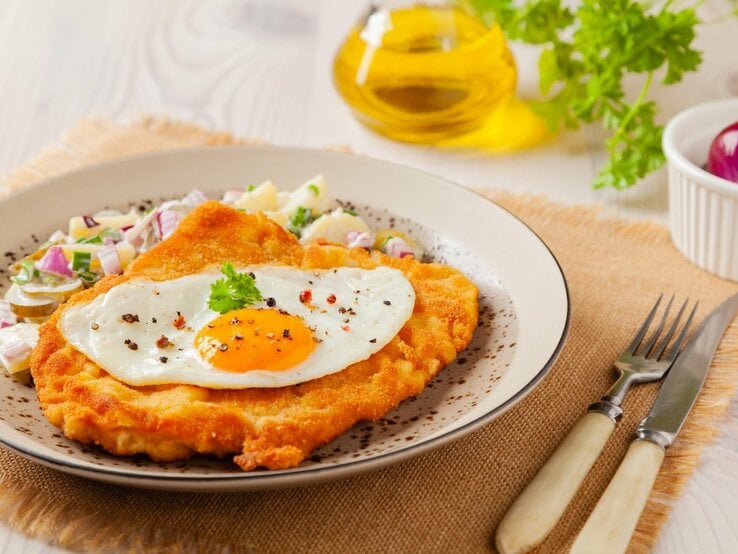 Ein appetitliches Wiener Schnitzel, gekrönt von einem Spiegelei mit einem glänzenden Dotter, liegt auf einem Teller mit fein gewürfelten Zwiebeln und Kräutern. Der Teller steht auf einer Stoffunterlage auf einem Holztisch. Im Hintergrund ist eine Flasche Olivenöl und ein Ramekin mit einer roten Zwiebel zu sehen. Ein Messer und eine Gabel mit Elfenbeingriffen liegen neben dem Teller