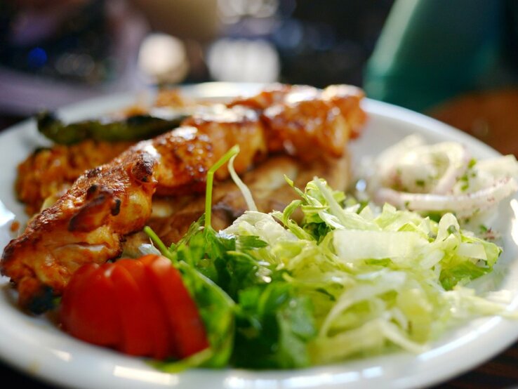 Auf einem weißen Teller sind türkische Hähnchenspieße serviert, begleitet von einem frischen, grünen Salat, einigen Tomatenscheiben und einer Portion Zwiebelringen in Joghurt-Dressing. Die Spieße sind appetitlich gebräunt und liegen im Vordergrund des Bildes, während der Salat und die Beilagen im Hintergrund verschwimmen, was auf eine geringe Tiefenschärfe hindeutet. 