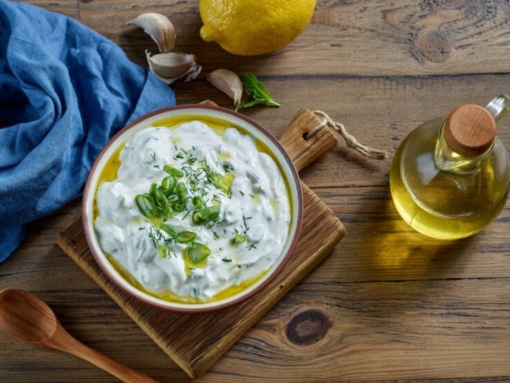 Ein farbenfrohes Bild zeigt eine Schale Çaçık, eine türkische Joghurtspeise, garniert mit geschnittenen Frühlingszwiebeln und Dill, auf einem Holzbrett. Neben der Schale befinden sich eine Zitrone, einige Knoblauchzehen, ein Bund frischer Minze und eine kleine Flasche Olivenöl mit einem Holzkorken. Eine blaue Stoffserviette liegt stilvoll gefaltet neben der Schale. Alle Elemente sind auf einer rustikalen Holzoberfläche arrangiert, die Wärme und Natürlichkeit ausstrahlt.