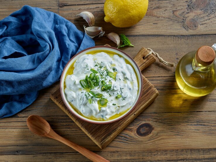 Ein farbenfrohes Bild zeigt eine Schale Çaçık, eine türkische Joghurtspeise, garniert mit geschnittenen Frühlingszwiebeln und Dill, auf einem Holzbrett. Neben der Schale befinden sich eine Zitrone, einige Knoblauchzehen, ein Bund frischer Minze und eine kleine Flasche Olivenöl mit einem Holzkorken. Eine blaue Stoffserviette liegt stilvoll gefaltet neben der Schale. Alle Elemente sind auf einer rustikalen Holzoberfläche arrangiert, die Wärme und Natürlichkeit ausstrahlt.