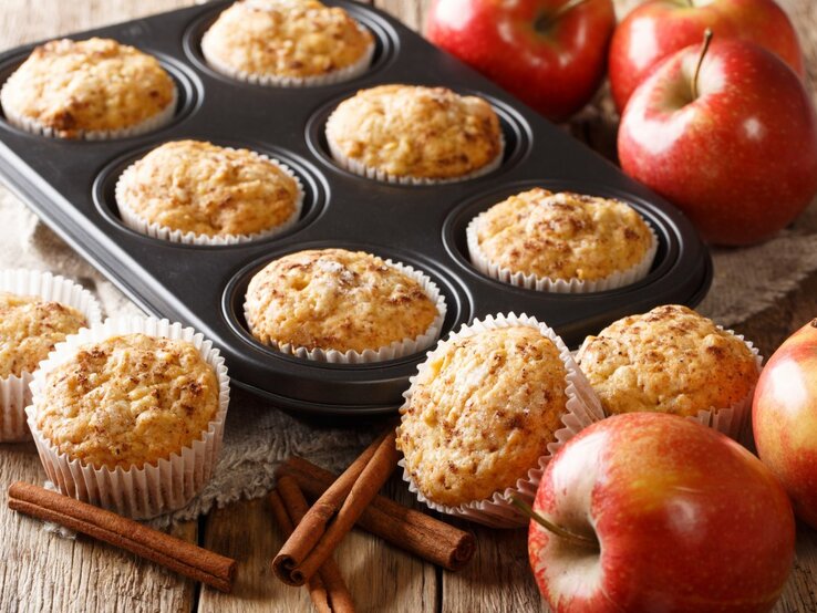 Frisch gebackene Apfelmuffins in einer Muffinform und auf einem Holztisch. Die Muffins sind goldbraun und sehen mit ihrer aufgegangenen, krümeligen Oberfläche appetitlich aus. Ein paar der Muffins sind noch in der schwarzen Muffinform, während andere auf dem Tisch neben der Form stehen, jeweils in weißen Papierförmchen. Rote Äpfel, Zimtstangen und ein Stück Sackleinen verleihen dem Bild eine rustikale und gemütliche Ausstrahlung. 