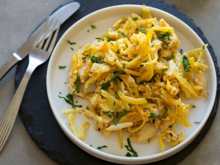 Alt-Text: Ein Teller mit Bacalhau à Brás, einem traditionellen portugiesischen Gericht aus zerpflücktem Stockfisch, der mit dünn geschnittenen, goldgelben Kartoffelstreifen und Eiern vermischt ist. Es ist garniert mit frischer, grüner Petersilie, die einen schönen Farbkontrast bildet. 