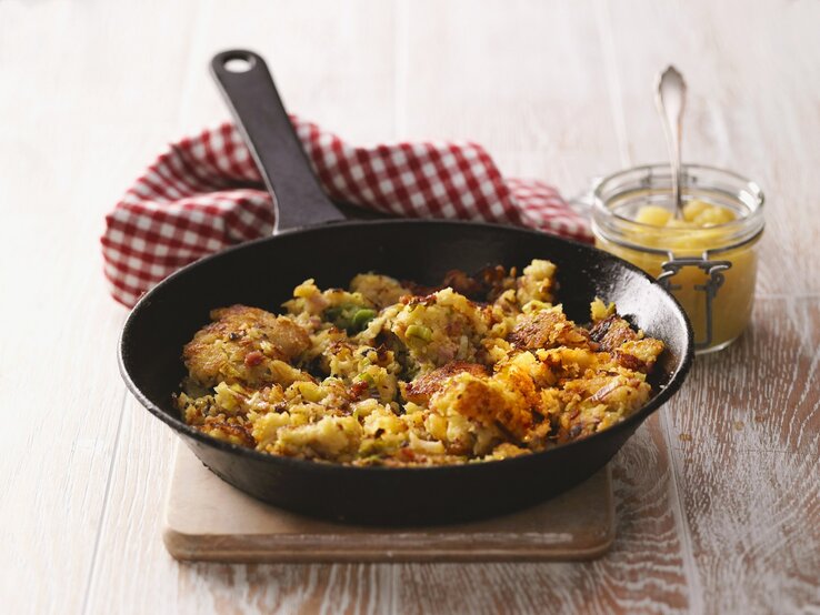 Eine Pfanne mit einem Pfannengericht mit Kartoffeln steht auf einer Holztischplatte. Daneben ein kleines Glas mit Apfelmus.