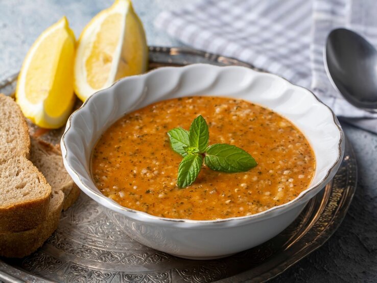 Ein Bild zeigt eine Schüssel mit Tarhana-Suppe, einer traditionellen türkischen Suppe, die reich an Gewürzen und Tomaten ist. Die Suppe hat eine leuchtend orangefarbene Textur mit sichtbaren Kräutern und Körnern, die eine rustikale und nahrhafte Mahlzeit suggerieren. Die Schüssel steht auf einem dekorativen Metalltablett und wird von zwei Zitronenspalten, einem Stapel Vollkornbrot und einem Löffel, der auf einem Stoffserviette liegt, flankiert. Ein Zweig frische Minze ziert die Mitte der Suppe, was einen farblichen Kontrast und einen Hinweis auf Frische gibt.