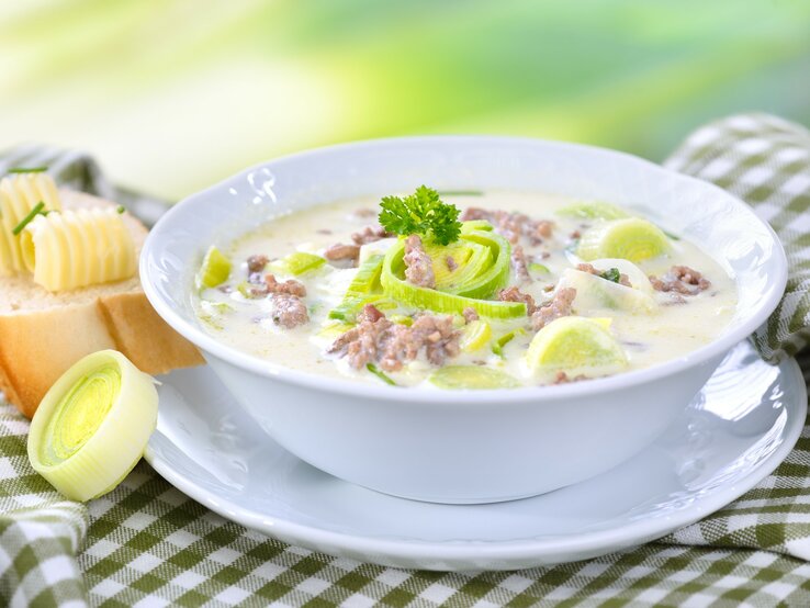 Das Bild zeigt eine Schüssel Suppe, die reichhaltig und cremig aussieht. Die Suppe ist mit Lauchringen garniert und es gibt sichtbare Stückchen von Hackfleisch. Neben der Suppenschüssel befindet sich eine Scheibe Brot mit einer Kugel Butter darauf. Das Brot und die Butter sehen frisch und einladend aus. Die Suppe scheint eine Käse-Lauch-Suppe zu sein, ein beliebtes Gericht in der deutschen Küche. 