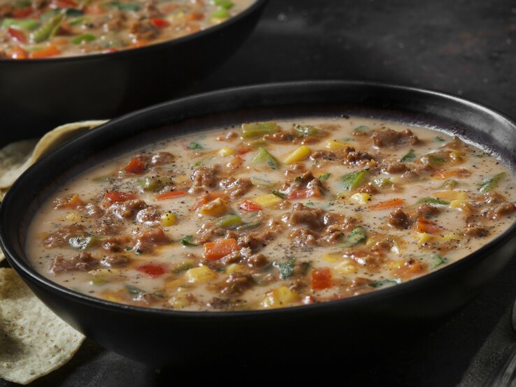 Das Bild zeigt eine herzhafte Schüssel Suppe, die mit Hackfleisch, Käse und verschiedenen Gemüsesorten wie Paprika, Zwiebeln und Mais zubereitet wurde. Die Suppe scheint cremig zu sein und ist großzügig mit schwarzen Pfeffer bestreut, was auf eine würzige Note hindeutet. 