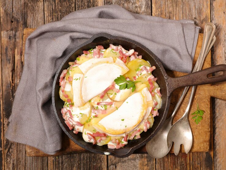 Das Bild zeigt eine Tartiflette, ein traditionelles französisches Gericht aus der Region Savoyen, das in einer gusseisernen Pfanne serviert wird. Es besteht aus Schichten von Kartoffeln, Speck und Zwiebeln, die reichlich mit Reblochon-Käse überbacken sind. Die oberste Käseschicht ist leicht gebräunt und geschmolzen, was auf eine kürzliche Zubereitung im Ofen hinweist. Garniert ist das Gericht mit frischen Kräutern, vermutlich Petersilie, die einen Farbtupfer geben. Das rustikale Holzbrett unter der Pfanne sowie das graue Stofftuch und das antike Besteck runden das Bild eines herzhaften, ländlichen Gerichts ab.