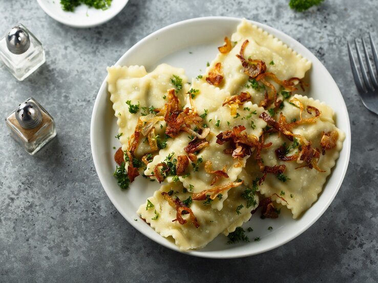 Mehrere Maultaschen mit Kräutern und Röstzwiebeln garniert auf weißem Teller und grauem Untergrund.