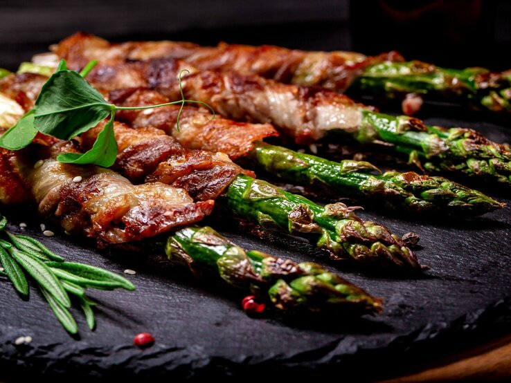 Appetitlicher Blick auf gebratenen Spargel in Speckmantel, fein gewürzt mit Kräutern auf Steinplatte.