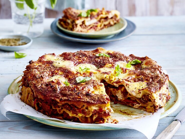 Eine angeschnittene Pizza-Lasagne auf einem gemusterten Teller auf einem blauen Untergrund. Im Hintergrund steht auf einem Teller noch ein Stück Pizza-Lasagne.