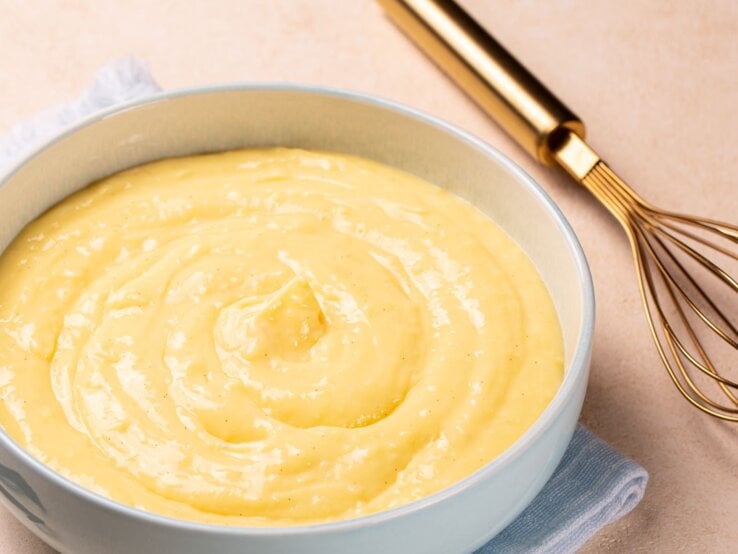 Low Carb Nachtisch: Cremiger Vanillepudding in einer hellblauen Schale auf einem hellblauen Tuch, daneben Vanilleschoten und ein goldener Schneebesen.