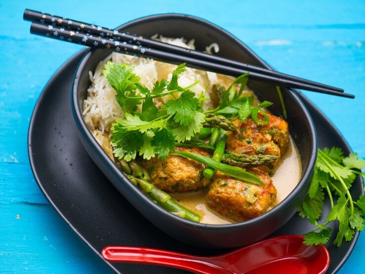Curry-Suppe-Spargel-Hackbaellchen Eine dunkle schale mit der Suppe und Reis auf blauem Untergrund.