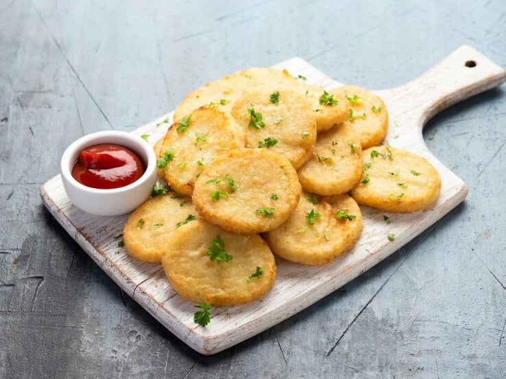 Mehrere flache Blumenkohlpuffer auf einem Holzbrett, daneben Ketchup. 