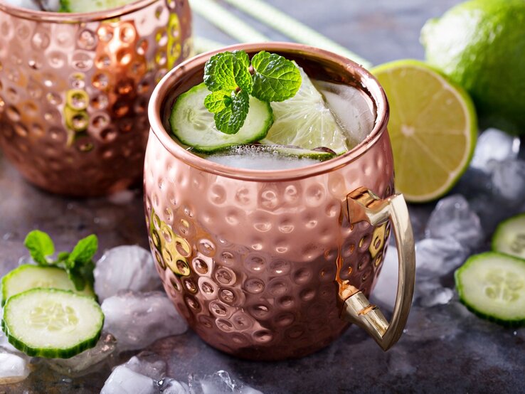Moscow Mule in einem gehämmerten Kupferbecher mit Eis, Limettenscheibe, Gurke und Minze auf einem Tisch mit Eiswürfeln und Limetten