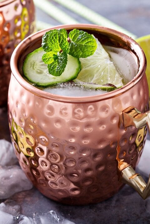 Moscow Mule in einem gehämmerten Kupferbecher mit Eis, Limettenscheibe, Gurke und Minze auf einem Tisch mit Eiswürfeln und Limetten