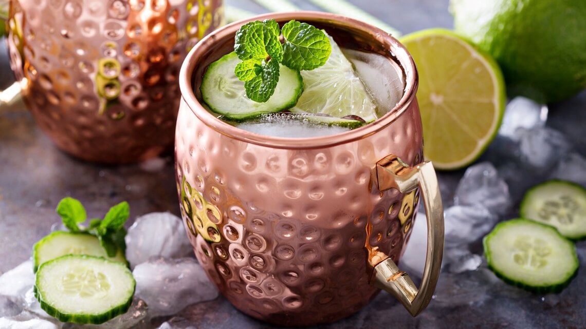 Moscow Mule in einem gehämmerten Kupferbecher mit Eis, Limettenscheibe, Gurke und Minze auf einem Tisch mit Eiswürfeln und Limetten
