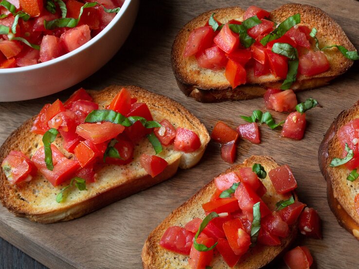 Knusprige Bruschetta mit frischen Tomatenwürfeln und Basilikumblättern auf Holzbrett neben einer weißen Schüssel.