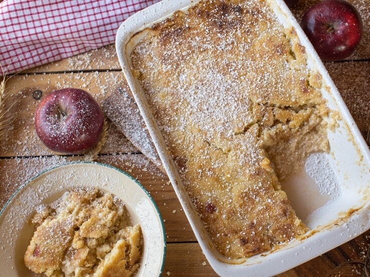 Frisch gebackener Apfelauflauf mit goldbrauner Kruste und Puderzucker in Auflaufform auf rustikalem Holztisch.