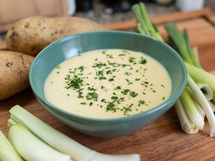 Vichysoisse Eine blaue Suppenschale mit der samtigen Suppe auf einem Holztisch. Drum herum Kartoffeln und Lauch.