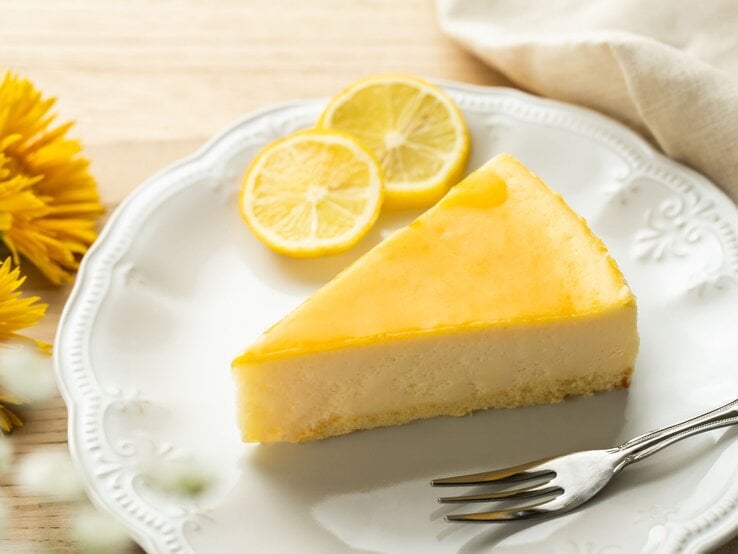 Ein Stück Zitronenkuchen mit glänzend gelber Glasur liegt auf einem verzierten weißen Teller, daneben Zitronenscheiben und gelbe Blüten.