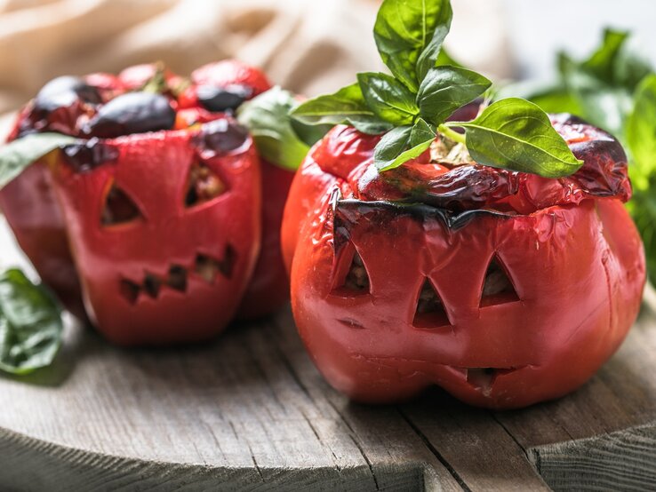 Zwei geschmorte Paprika im Halloween-Look auf einem Holzbrett mit Basilikum dekoriert. Alles von der Seite und recht nah fotografiert.