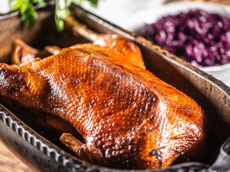 Eine goldbraune gebackene Gans in einer tiefen Backform auf einem Holzschneidebrett und hellem Küchentisch. Im Hintergrund sind unscharf Rotkohl und die Brezeln zu erkennen. | © Shutterstock/ Marian Weyo