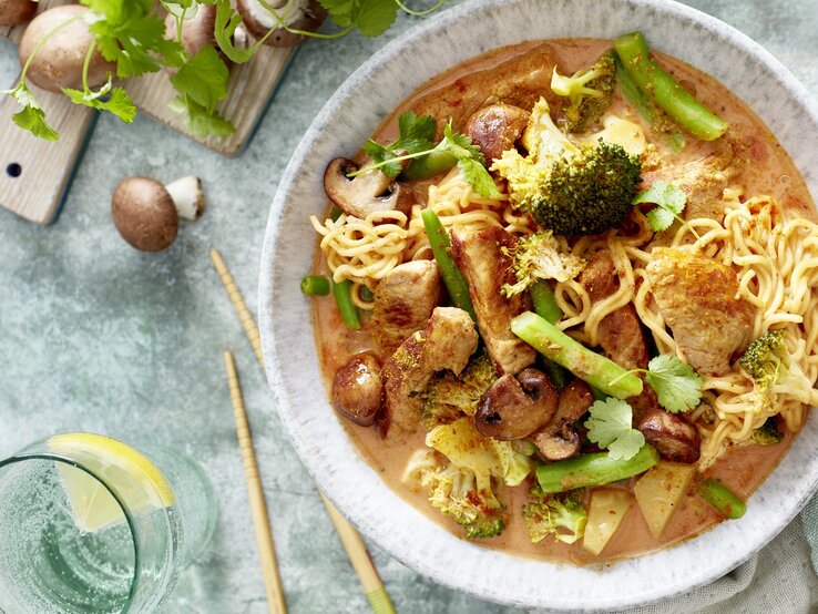 Ein helle Teller mit dem Thai-Curry auf einem hellblauem Untergrund mit grünem Gemüse und Pilzen daneben. Alles direkt von oben fotografiert.