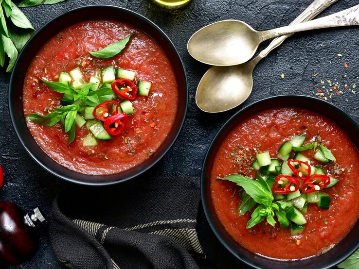 Zwei runde Schalen mit der spanischen Gemüsesuppe Gazpacho Andaluz / zwei Löffel / frischer Knoblauch / Kräuter / Pfeffermühle /Tuch / eine Tomate / Draufsicht