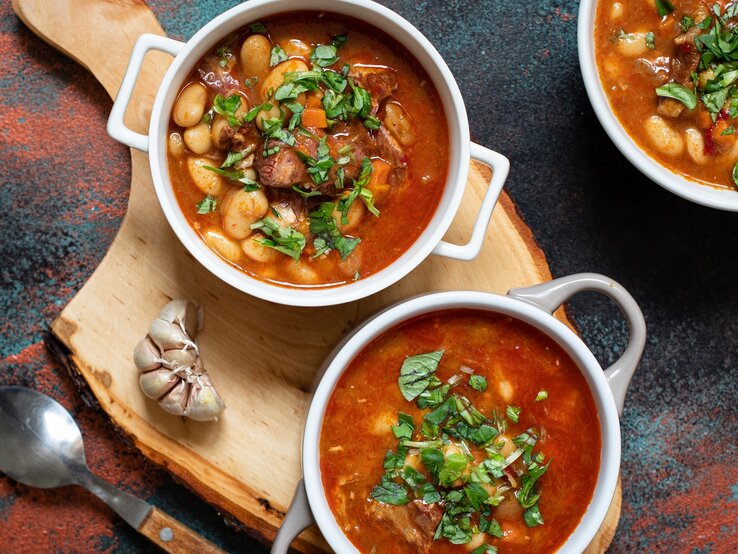 Serbische Bohnensuppe in drei runden Schalen aus Porzellan / Holzbrett / frischer Knoblauch / Suppenlöffel / Draufsicht