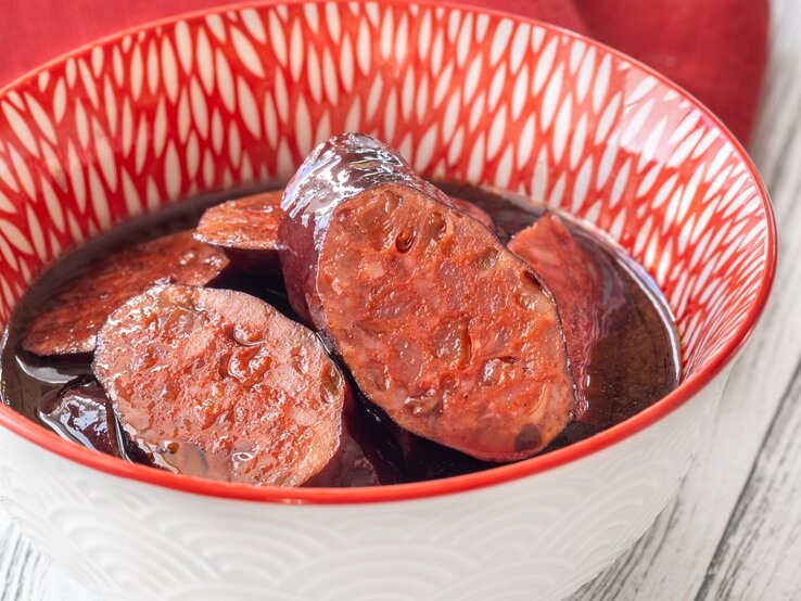 Eine weiß-rote Schale mit dem Tapasgericht auf einem hellen Holztisch. Alles von schräg oben fotografiert.