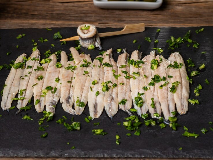 Die in Essigsud eingelegten Sardellenfilets auf einer schwarzen Schieferplatte. Alles von schräg oben fotografiert.