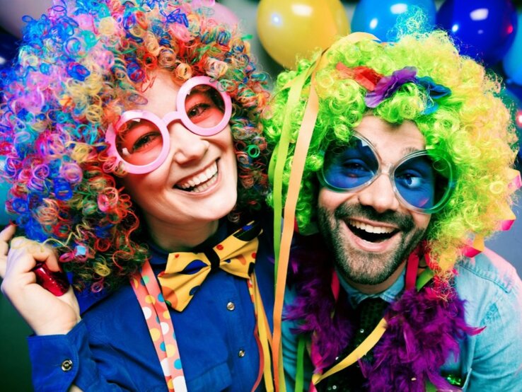  Ein fröhliches Paar, verkleidet in bunten Karnevalskostümen, mit übergroßen Brillen, farbenfrohen Perücken und festlichen Accessoires. Die Frau trägt eine blaue Jacke und eine pinkfarbene Brille, während der Mann eine bunte Fliege, eine lila Federboa und eine blaue Brille mit großen Gläsern trägt. Beide strahlen Freude und Ausgelassenheit aus, umgeben von Luftballons im Hintergrund, was eine ausgelassene Karnevalsstimmung vermittelt.