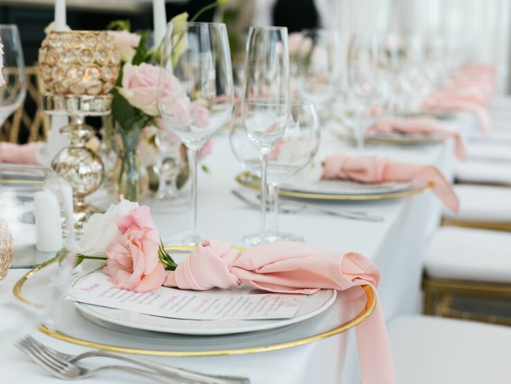 Das Bild zeigt eine elegant gedeckte Tafel, die für eine formelle Veranstaltung oder Feier vorbereitet wurde. Im Vordergrund sieht man einen Tisch mit weißer Tischdecke, der mit verschiedenen Elementen für ein feines Dining-Erlebnis eingedeckt ist.  Es sind mehrere Gedecke zu erkennen, jedes mit einem Teller, der von goldfarbenen Besteckteilen flankiert wird. Über jedem Teller liegt eine rosafarbene Serviette, die kunstvoll gefaltet und drapiert ist, sodass sie über den Rand des Tellers fließt und eine elegante und weiche Linie bildet, die zur Ästhetik des Tisches beiträgt. Es ist möglich, dass die Serviette in einer einfachen, aber stilvollen Faltung wie einer Rolle oder einer einfachen Fächerfaltung gelegt wurde, welche die Serviette dekorativ hervorhebt, ohne zu komplex zu sein.  Auf dem Teller liegt zudem eine Menükarte, die vermutlich die Speisenauswahl für das Event zeigt. Dies deutet darauf hin, dass es sich um ein vorab geplantes Menü handelt, was typisch für Hochzeiten oder Bankette ist.  Die Tischdekoration umfasst auch florale Elemente – in diesem Fall zarte rosa Blumen, die wahrscheinlich als Echinochea oder Pfingstrosen identifiziert werden könnten – und ergänzende Kerzenhalter, die eine warme und einladende Atmosphäre schaffen. Die Gläser, die für Wasser, Wein und möglicherweise Champagner oder einen anderen Aperitif gedacht sind, stehen in einer ordentlichen Reihe hinter dem Teller und vervollständigen jedes Gedeck.