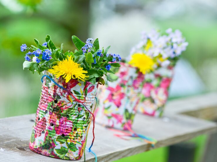 Zwei handgefertigte Blumentöpfe aus recycelten Konservendosen, verziert mit bunten, floralen Mustern und umwickelt mit farbenfroher Schnur, stehen auf einem Holzbrett im Freien. Die Dosen sind bepflanzt mit einer Auswahl an Wildblumen, einschließlich gelber Löwenzahn und blauer Vergissmeinnicht, die fröhlich in die Höhe ragen.
