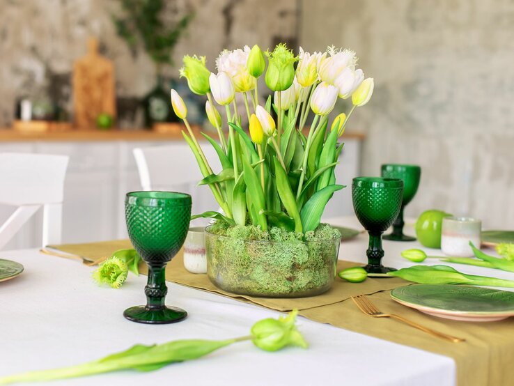 Ein frischer Strauß weißer und gelber Tulpen, arrangiert in einer durchsichtigen Glasvase mit grüner Moosfüllung, steht als Tischdekoration in einem hellen Esszimmer. Die Tischdekoration ist farblich aufeinander abgestimmt, mit grünen Glasweingläsern, hellgrünen Tellern und goldfarbenen Besteck auf einem olivgrünen Tischläufer. Im Hintergrund ist die moderne, helle Küche mit warmen Holzelementen zu erkennen, die eine einladende und frühlingshafte Atmosphäre schafft.