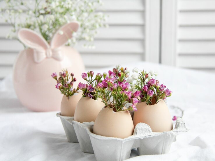 Ein frühlingshaftes Bastelarrangement mit Eierschalen, die als Miniaturpflanzgefäße für zierliche rosa Blumen dienen. Die Eierschalen sind in einer Eierkarton-Halbschale angeordnet, die auf einem weißen Tischtuch steht. Im Hintergrund ist eine zarte, rosafarbene Keramikvase in Hasenform zu sehen, die das Osterthema unterstreicht und der Szene einen Hauch von Festlichkeit verleiht.
