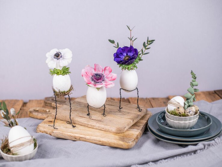  Drei dekorative Eier auf Drahtbeinen mit aufgesetzten Blumenköpfen – ein weißes mit einer Anemone, ein zweites mit einer rosa Mohnblume und ein drittes mit einer dunkelvioletten Anemone – arrangiert auf einem hölzernen Schneidebrett. Auf dem Brett befinden sich auch weitere dekorierte Eier in einer kleinen Schüssel auf einem Stapel von Keramiktellern. Die Szene ist auf einem grauen Tuch mit lila Wand im Hintergrund drapiert, was einen kreativen Basteltrend mit Draht und Naturmaterialien darstellt.