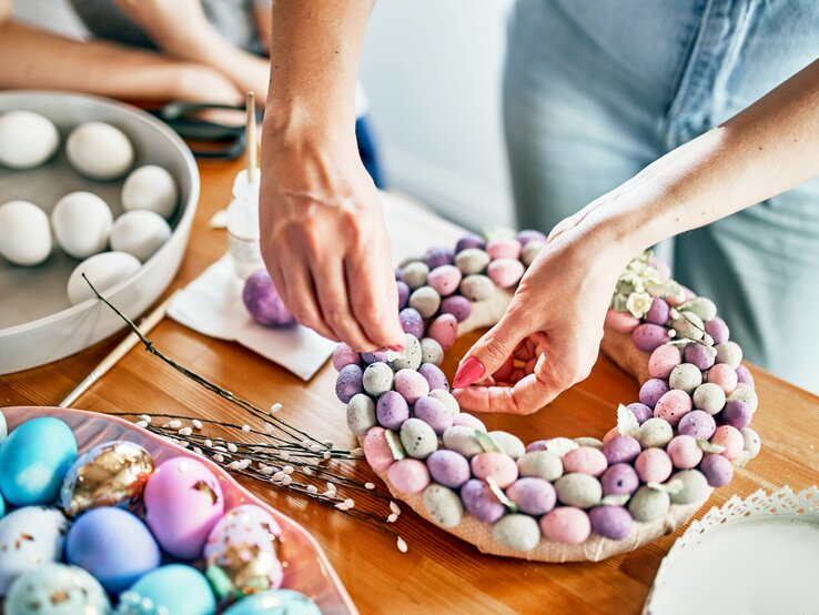 Das Bild zeigt eine Person, die an einem Bastelprojekt mit Ostereiern arbeitet. Sie erstellt anscheinend einen Kranz, der mit vielen kleinen Eiern in verschiedenen Pastellfarben wie Lila, Rosa und Blau verziert ist. Die Eier sehen aus, als könnten sie aus Schaumstoff oder einem ähnlichen Material gefertigt sein, und sind zu einer runden Form angeordnet. Auf dem Tisch sind auch andere Bastelmaterialien zu sehen, darunter eine Schale mit echten, weiß gefärbten Eiern, ein Teller mit zusätzlichen dekorativen Eiern in einer Mischung aus pastellfarbenen und metallischen Tönen sowie einige Zweige, die wahrscheinlich auch als Teil des Dekorationsprojekts dienen. Die Szene wirkt fröhlich und festlich, passend zu einer Osterbastelaktivität.