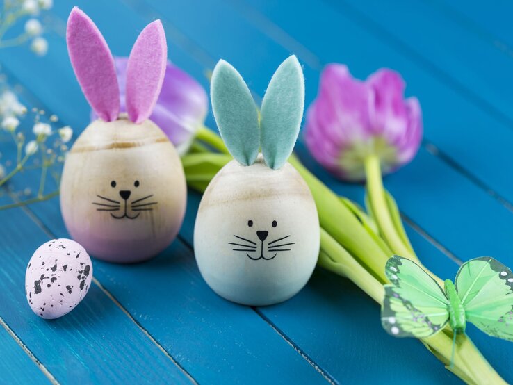  Zwei kreative Osterhasen-Dekorationen aus Eiern mit aufgemalten Gesichtern und Ohren aus farbigem Filz auf einem leuchtend blauen Holzhintergrund, begleitet von einem zarten, gesprenkelten Osterei, frischen Tulpen und einem Schmetterlingsornament auf grünen Stängeln, was ein fröhliches und buntes Ambiente für Ostern schafft.