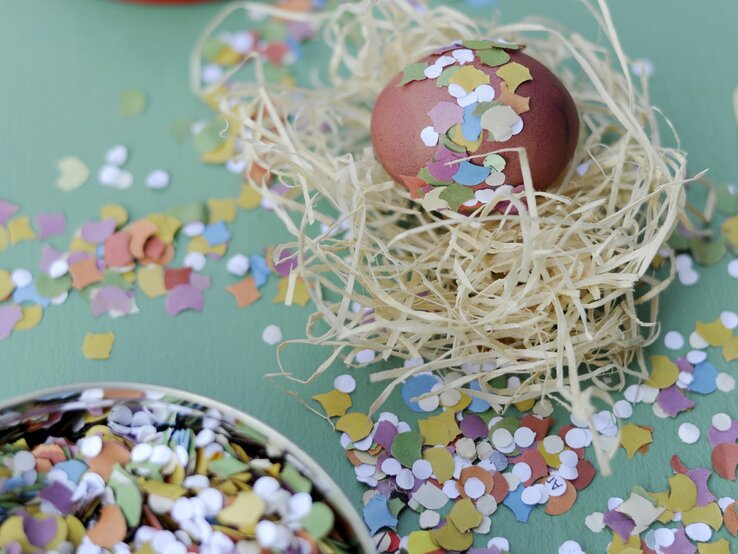  Das Bild zeigt ein sorgfältig dekoriertes Osterei, das in einem Nest aus natürlich aussehendem Stroh platziert ist. Das Ei ist mit bunten Konfettistücken verziert, die auf seiner Oberfläche haften und ihm ein festliches Aussehen verleihen.