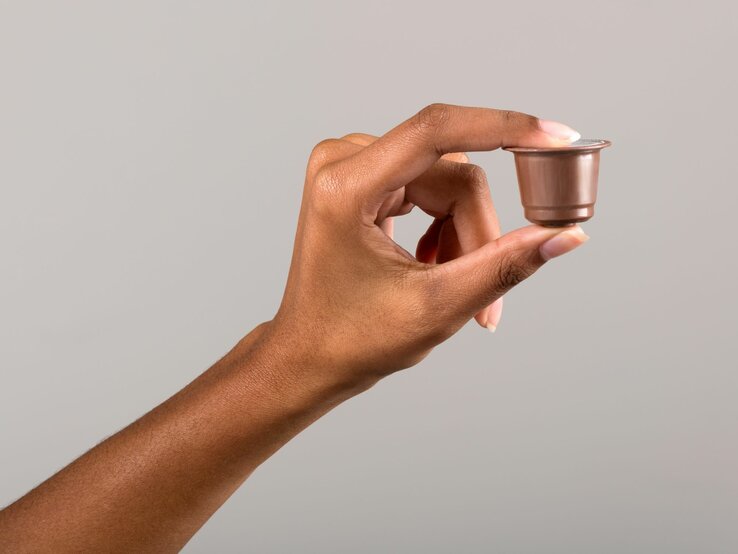  Ein Bild einer Hand, die eine gebrauchte Kaffeekapsel zwischen Daumen und Zeigefinger hält. Der Hintergrund ist einfarbig und neutral, um den Fokus auf die Kapsel zu lenken.