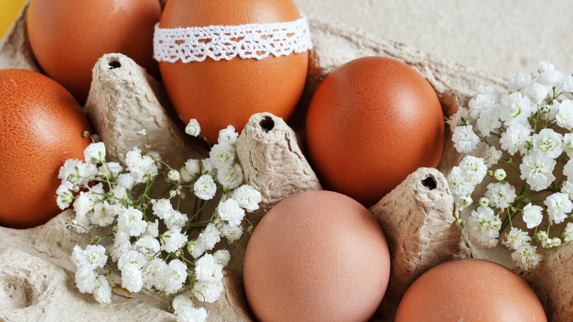 Ein Eierkarton aus Pappe enthält mehrere braune Eier. Eines der Eier ist mit einer dekorativen weißen Spitzenborte umwickelt. Zwischen den Eiern sind kleine Zweige mit weißen Blüten drapiert, was dem Ganzen eine frühlingshafte und österliche Atmosphäre verleiht. Der Hintergrund ist gelb, was den warmen, fröhlichen Charakter der Komposition betont.